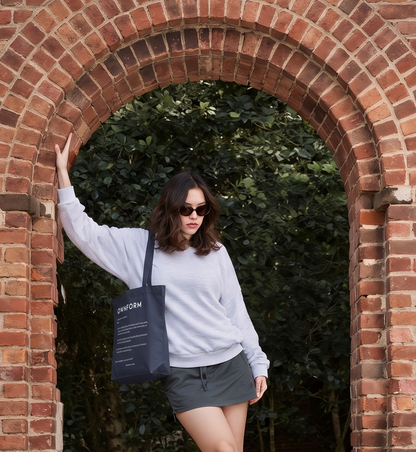A woman wearing a light gray sweatshirt and dark gray shorts stands in front of a red brick archway, carrying a black tote bag with white text that reads 'OWNFORM'.